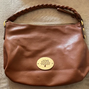 Brun axelväska från Mulberry i skinn - Säljer en snygg brun axelväska från Mulberry i äkta skinn med flätat handtag och guldfärgad logoplatta framtill. Väskan har dragkedja upptill och ett rymligt innerfack med mönstrat foder. Perfekt för dig som vill ha en klassisk och stilren väska med lyxig känsla.