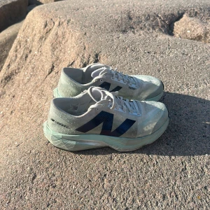 New Balance FuelCell Rebel v2 löparskor  - Lätta och sportiga New Balance FuelCell Rebel v2 sneakers i ljusgrått med mintgrön sula och stora mörkblå N-loggor på sidorna. Ovandelen är i ventilerande syntetmaterial och skorna har snörning samt platt sula. Perfekta för löpning eller träning. Nypris: 2300kr