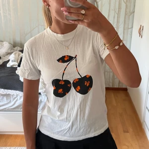 Vit t-shirt med körsbärstryck från Ganni - INTRESSEKOLL🥰 Vit t-shirt från GANNI med svart och orange körsbärstryck framtill. T-shirten har rund hals, korta ärmar och normal passform. Trycket har små orange blommor på svarta körsbär. Perfekt för en chill och lekfull look. (Skriv till mig vid intresse)❤️