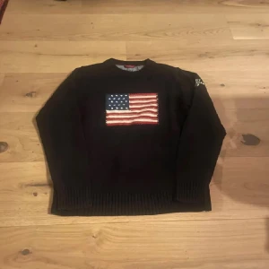 Svart stickad Gant tröja med USA-flagga - Svart stickad tröja från Gant i 100% bomull. På bröstet sitter en stor, broderad amerikansk flagga i rött, vitt och blått. Tröjan har rund hals och långa ärmar med ribbade muddar för en klassisk look.
