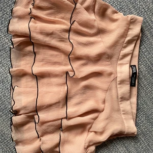 Volangkjol från Zara - Supersöt rosa/beige kortkjol från Zara med flera lager volanger och svarta kanter. Tillverkad i ett lätt och luftigt material som ger snyggt fall💓