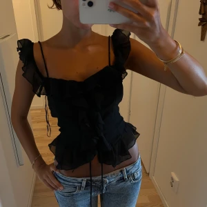 Svart volangig blus med smala axelband - Supersöt svart topp med volanger och smala axelband. Toppen har en feminin v-ringning, volangdetaljer längs axlar och framtill samt en lätt transparent känsla. 