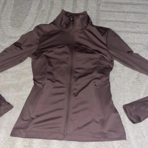 Brun sportig vindjacka med dragkedja - Slimmad brun vindjacka med hög krage och hel dragkedja framtill. Jackan har långa ärmar och två fickor med dragkedja på sidorna. Perfekt för dig som gillar en sportig och stilren look.