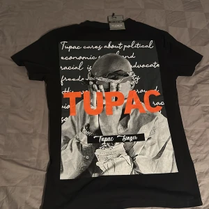 Svart Tupac t-shirt från Kauma - Svart t-shirt med stor Tupac-print på ryggen och mindre tryck framtill. Snyggt motiv i svartvitt med orange text och coola detaljer. Klassisk passform och rund hals. Perfekt för dig som gillar streetstyle och hiphop-vibe.