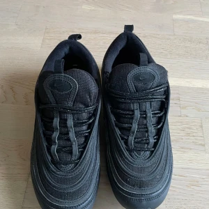 Svarta Nike Air Max 97 sneakers - Säljer ett par helsvarta Nike Air Max 97 sneakers med ikoniska vågformade detaljer och synlig Air-sula. Skorna har snörning, mesh och syntetmaterial i ovandelen samt dragflikar både fram och bak. Perfekta för dig som gillar en clean och sportig look.