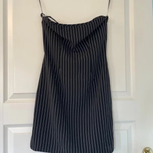 Svart kortklänning med smala ränder - Snygg svart kortklänning med tunna vita ränder och smala axelband. Klänningen har en lätt draperad halsringning och snörning i ryggen för extra detalj. Perfekt för dig som gillar en stilren och trendig look. Sitter GALET snyggt på! Endast använd en gång. Stretchiga, strl s