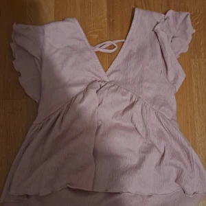 Ljuslila/rosa omlott blus med volangärm - Supersöt ljuslila/rosa blus med omlott och v-ringning. Toppen har korta volangärmar och croppad passform, samt knyt i ryggen. Perfekt för vår och sommar när du vill ha en chill men ändå söt look.