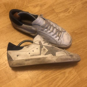 Golden goose skor - Golden Goose sneakers i vitt skinn med grå stjärndetalj på sidan och mörkblå häl. Klassisk låg modell med snörning och platt sula. Snyggt slitna detaljer och bruna yttersulor. Perfekta för dig som gillar streetstyle med edge. Endast box medföljer. Storlek 44