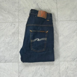 Mörkblå jeans från Nudie - Snygga mörkblå jeans från Nudie i modellen Thin Finn. Klassisk femficksdesign, orange kontrastsömmar och ikonisk våg på bakfickan. Tight passform och tillverkade i stretchig bomullsdenim för en bekväm känsla.