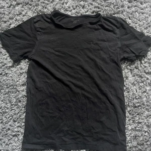 Svart t-shirt från Calvin Klein - En stilren svart t-shirt från Calvin Klein med diskret 'ck'-brodyr på bröstet. Klassisk passform, rund hals och korta ärmar. Tillverkad i mjuk bomull som känns skön mot huden. Perfekt för en clean och enkel look.
