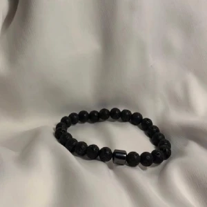 Svart pärlarmband med lavasten - Stilrent armband med svarta runda lavastenpärlor och en metallisk detalj. Elastiskt band gör det enkelt att ta på och av. Perfekt accessoar för en cool och minimalistisk look.
