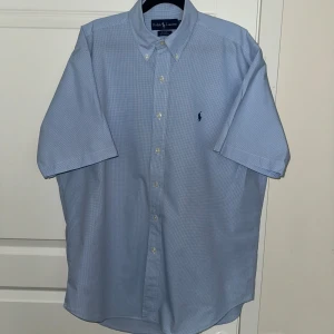 Blå rutig skjorta från Ralph Lauren - Snygg blå rutig skjorta från Ralph Lauren i 100% bomull. Skjortan har korta ärmar, klassisk button-down krage och knappar framtill. Den ikoniska Polo-loggan är broderad på bröstet. Perfekt för dig som gillar stilrena och klassiska plagg.