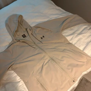 Beige vindjacka från Under Armour - Snygg beige vindjacka från Under Armour med huva och dragkedja hela vägen. Jackan har svarta detaljer vid snörningen och logga på bröstet. Perfekt för blåsiga dagar och har praktiska fickor framtill. Lätt och smidig att bära med sig.
