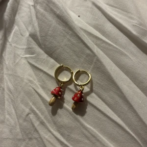 Guldiga örhängen med svampberlocker - Supergulliga örhängen i guld med små hängande svampberlocker. Svamparna har röd hatt med vita prickar och beige fot, vilket ger en lekfull och trendig look. Perfekta för dig som vill sticka ut med något unikt och färgglatt i öronen. Använt ett fåtal gånger men bra skick! Från fable england