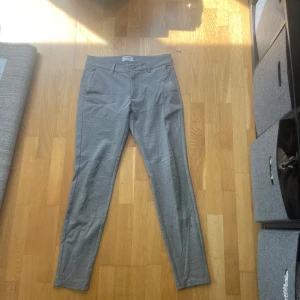 Grå kostymbyxor från Only & Sons - Storlek 30/34 Snygga grå kostymbyxor från Only & Sons med klassisk passform och raka ben. Byxorna har bälteshällor, sidfickor och bakfickor med knapp. Perfekta för en clean och stilren look. Materialet är mjukt och känns bekvämt hela dagen.