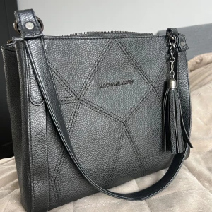 Väska från Michael Kors - Snygg svart axelväska från Michael Kors i präglat skinn med geometriska sömmar och en dekorativ tofs. Väskan har dragkedja upptill och ett stilrent, modernt uttryck. Perfekt storlek för det viktigaste och enkel att bära över axeln.