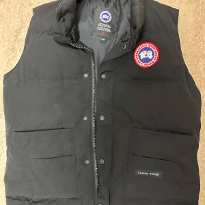 Svart dunväst från Canada Goose - Svart dunväst från Canada Goose med klassisk logga på bröstet. Västen har två stora fickor framtill, dragkedja och tryckknappar. Perfekt för lager-på-lager och riktigt varm. Stilren och enkel design som funkar till det mesta. Pris kan diskuteras men kan ej fraktas!