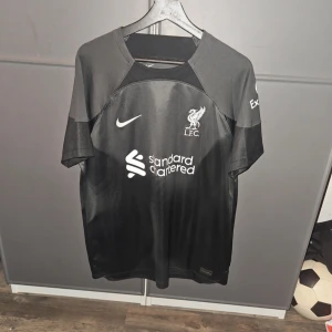 Svart Liverpool Nike fotbollströja L Äkta - Snygg svart Liverpool FC fotbollströja från Nike i storlek L. Tröjan har korta ärmar, rund hals och är tillverkad i lätt och ventilerande Dri-FIT polyester. Tryck med klubbmärke, Nike-logga och sponsor på bröstet. Perfekt för dig som älskar fotboll och vill representera LFC.