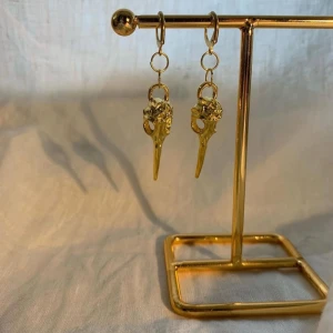 Guldiga örhängen med fågelskalle - Unika örhängen i guldton med detaljerade hängen formade som fågelskallar. De har en cool och edgy vibe, perfekta för dig som vill sticka ut. Hängena är detaljerade och ger en riktigt snygg look.
