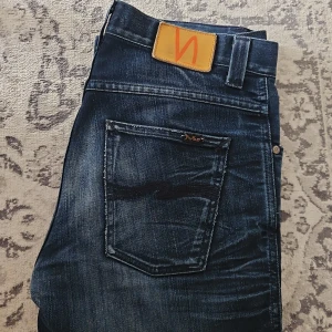 Nudie Jeans Slim Jim Black n Blue 30/32 - Snygga Nudie Jeans Slim Jim i tvätten Black n Blue. Jeansen har en mörkblå tvätt med slitningar, klassisk femficksdesign och dragkedja. Slim fit-modell i bomull med snygga detaljer på bakfickan och läderpatch i midjan. Eventuellt upplaga efter 170cm lång användare. 