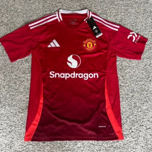 Manchester United tröja  - Röd Manchester United fotbollströja från Adidas i slim fit. Tröjan har vita detaljer, klubbmärke på bröstet och Snapdragon-logga framtill. Tillverkad i lätt och ventilerande material med klassiska Adidas-ränder på axlarna. Perfekt för dig som älskar fotboll och vill sticka ut på planen.