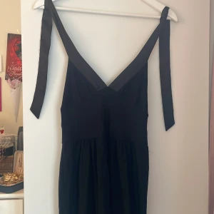 Svart klänning med knytband - Stilren svart klänning med v-ringning och breda axelband som knyts upptill. Modellen är midilång med lätt rynkad kjol och markerad midja. Perfekt för dig som gillar enkel men snygg design. Materialet känns mjukt och följsamt.