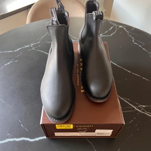 Svarta chelsea boots från R.M. Williams - Klassiska svarta chelsea boots från R.M. Williams i slätt skinn. Snygg minimalistisk design med rund tå, resår på sidorna och dragflikar med logga. Perfekta till jeans eller kostymbyxor för en clean look.