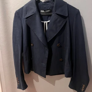 Mörkblå trenchcoat från Zara - Säljer en mörkblå trenchcoat från Zara med dubbelknäppning och breda slag. Jackan har snygga knappar framtill och vid ärmsluten, samt en croppad passform som ger en modern look. Perfekt att slänga över en t-shirt för en cool stil.