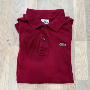 Vinröd pikétröja från Lacoste - Snygg vinröd pikétröja från Lacoste med klassisk krage och två knappar framtill. Ikoniska gröna krokodilloggan broderad på bröstet. Tillverkad i bomull, perfekt för en clean och sportig look. Kortärmad modell som passar året runt.