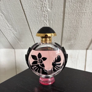 Paco Rabanne Olympea parfymflaska - Snygg parfymflaska från Paco Rabanne Olympea med ljusrosa doft, rund form och dekorativa svarta blommor och blad i glaset. Korken är svart med gulddetaljer och flaskan står på en rosa botten. Perfekt som statement på sminkbordet.