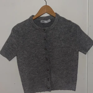 Grå stickad kofta från Zara - Kortärmad grå stickad kofta från Zara med rund halsringning och knappar framtill. Mjuk och mysig känsla, perfekt att slänga över en topp. Ribbstickade detaljer vid ärmslut och nederkant ger en snygg finish.