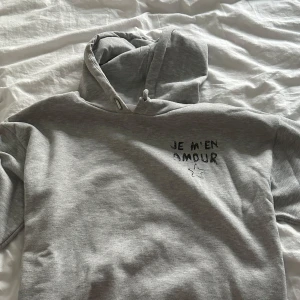 Grå hoodie från Gina Tricot - Grå hoodie från Gina Tricot med svart texttryck 'JE M'EN AMOUR' och en stjärna på bröstet. Mjuk insida, huva med snörning och klassisk passform. Perfekt för dig som gillar chill och enkel stil.
