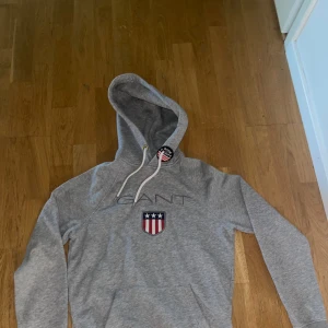 Grå hoodie från GANT - Snygg grå hoodie från GANT i bomull med broderad logga och amerikanskt inspirerat märke på bröstet. Tröjan har huva med vit snörning och en extra broderad detalj vid snöret. Storlek xs. Har används några gånger men är i bra skick. Skriv om du har frågor💕. Pris kan diskuteras.