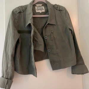 Grön overshirt från Twist & Tango - Snygg militärgrön overshirt från Twist & Tango med knappar, dragkedjedetaljer och axelklaffar. Jackan har långa ärmar och en rak, lite kortare passform. Perfekt att slänga över en hoodie eller t-shirt för en cool look.