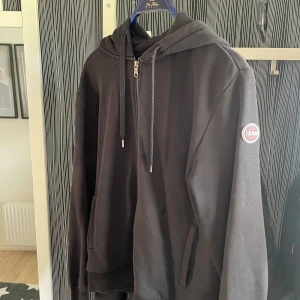 Svart hoodie från Colmar med huva - Svart hoodie från Colmar med huva, dragsko och half zip framtill. Klassisk och enkel design med Colmar-logga på ärmen. Perfekt för en chill och avslappnad stil. Mjuk bomullskänsla och rymliga fickor framtill.