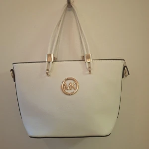 Vit handväska från Michael Kors - Snygg vit handväska från Michael Kors med guldiga detaljer och MK-logga framtill. Väskan har två rejäla handtag, dragkedja baktill och stilren form. Perfekt för dig som vill ha en klassisk och trendig accessoar.