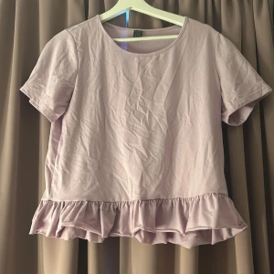 Ljuslila volangtopp med kort ärm - Supersöt ljuslila topp med volangkant nertill och korta ärmar. Toppen har en rund halsringning och är gjord i mjuk bomull, perfekt för en chill och feminin look. Enkel att styla med jeans eller kjol för en söt vibe.