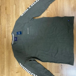Grå långärmad tröja Abercrombie - Snygg grå långärmad tröja från Abercrombie & Fitch med tryckt logga längs ena ärmen och broderad logga på bröstet. Tröjan har en bröstficka och rund halsringning. Perfekt för en chill och stilren look.