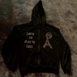 Svart rhinestone hoodie från bing lucid  - Svart hoodiejacka från bing lucid i storlek S. Framsidan har strasstenar som bildar texten 'paris au clair de lune' och ett motiv av Eiffeltornet och månen. Jackan har dragkedja, huva och två fickor framtill. Perfekt för dig som gillar statement-plagg.