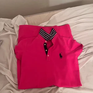 Intressekoll av Rosa pikéklänning från Ralph Lauren - Snygg rosa pikéklänning från Ralph Lauren med klassisk krage och knappar framtill. Klänningen har korta ärmar, marinblå logga på bröstet och randig detalj på insidan av kragen. Perfekt för en sportig och fräsch look. Orginal pris 2600kr.