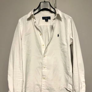 Vit skjorta från Ralph Lauren - Klassisk vit skjorta från Ralph Lauren med broderad marinblå logga på bröstet. Skjortan har lång ärm, knappar framtill och är i bomull med custom fit-passform. Perfekt för en clean och stilren look. Sitter som xs.