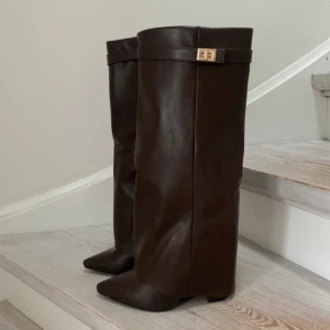 Bruna höga stövlar med gulddetalj - Snygga höga stövlar i mörkbrunt skinn med spetsig tå och guldfärgad spänndetalj upptill. Stövlarna har en elegant och stilren look med låg klack och slät yta. 