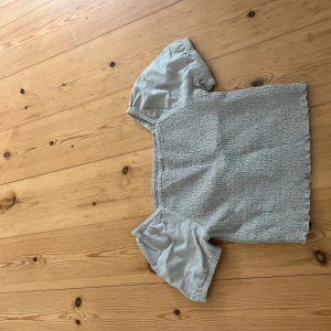 Ljusgrön blus från Kappahl - Supersöt ljusgrön blus från Kappahl med puffiga korta ärmar och smockad kropp. Toppen har en fyrkantig halsringning och är gjord i mjuk bomull, perfekt för en chill och trendig look.