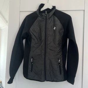 Svart vindjacka från Cross Sportswear - Snygg svart vindjacka från Cross Sportswear i storlek S. Jackan har hel dragkedja, två fickor med dragkedja och sportig design. Materialet är lätt och smidigt, perfekt för utomhusaktiviteter. Logga på bröstet och stilren look.