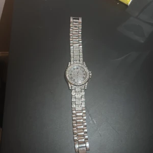 Iced out silverfärgad klocka - Säljer en riktigt blingad klocka i silverfärg med massor av glittrande stenar på både armband och urtavla. Klockan har romerska siffror och en chunky länkdesign som verkligen sticker ut. Perfekt för dig som vill ha en statement-accessoar som syns.