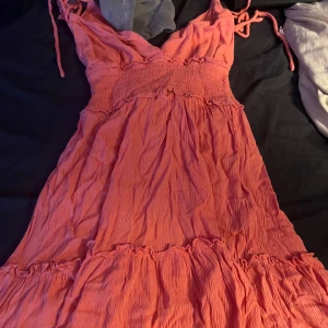 Korallrosa sommarklänning med volang - Superfin korallrosa klänning med smock i midjan, volangdetaljer och tunna axelband som knyts. Lätt och luftig modell med v-ringning och skrynklat tyg, perfekt för varma dagar.