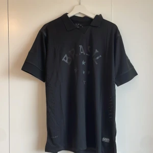 Svart Brasil Nike fotbollströja XL - Snygg svart Brasil fotbollströja från Nike i storlek XL. Tröjan har korta ärmar, diskret Brasil-tryck och stjärnor på bröstet samt ventilerande meshpaneler på sidorna. Tillverkad i lätt och tekniskt Dri-FIT-material som andas. Perfekt för dig som gillar stilren sportdesign.