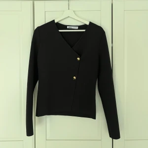 Svart omlottkofta från Zara - Stilren svart omlottkofta från Zara med v-ringning och två snygga guldfärgade knappar framtill. Koftan har lång ärm och är tillverkad i ett mjukt, ribbat material som ger en skön passform. Perfekt för dig som gillar minimalistisk och trendig stil.