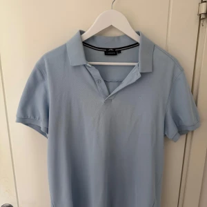 J.Lindeberg pikétröja – ljusblå, strl M - Snygg och klassisk pikétröja från J.Lindeberg i en fräsch ljusblå färg. Modellen är i storlek Medium och passar perfekt för både vardag och lite mer uppklädda tillfällen. Mjukt och bekvämt material med diskret JL-logga på bröstet.