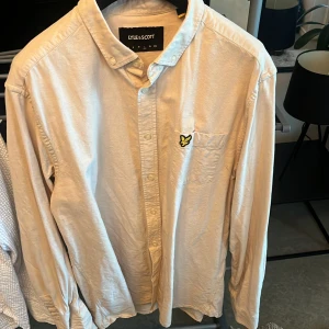 Beige skjorta från Lyle & Scott - Snygg beige skjorta från Lyle & Scott med klassisk krage och knappar hela vägen. Bröstficka med den ikoniska gula örnloggan. Skjortan är långärmad och har en clean, stilren look som passar till många outfits.